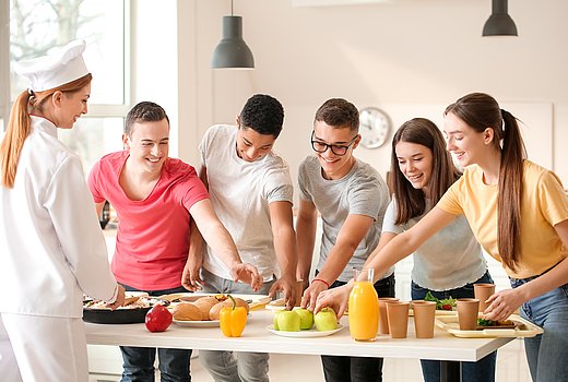 Auf einem weißen Tisch sind Speisen, Obst und Gemüse präsentiert. Davor stehen fünf junge Erwachsene mit ihren Tabletts, auf denen Essen steht. Sie greifen alle gemeinsam nach einem Teller mit grünen Äpfeln. 