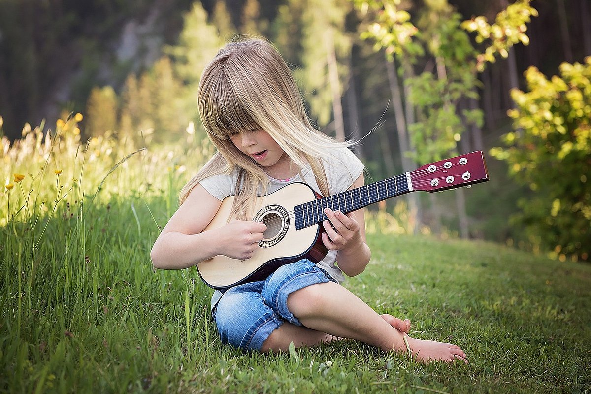 Kleines Mädchen mit langen blonden Haaren sitzt auf einer Wiese am Wald und spielt auf einer Spielzeug-Gitarre. 