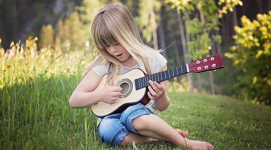 Kleines Mädchen mit langen blonden Haaren sitzt auf einer Wiese am Wald und spielt auf einer Spielzeug-Gitarre. 