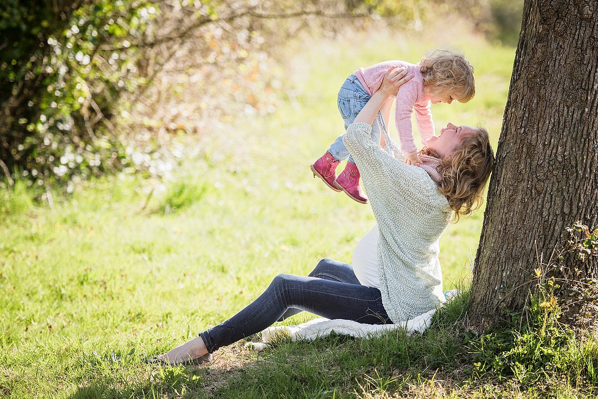 EIne schwangere Frau sitzt auf einer Wiese unter einem Baum und hält ein Kleinkind auf dem Arm in die Luft. Beide lachen.