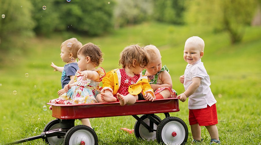 Eine Gruppe von fünf Kleinkindern sitzt und steht um einen roten Bollerwagen auf einer Wiese herum.