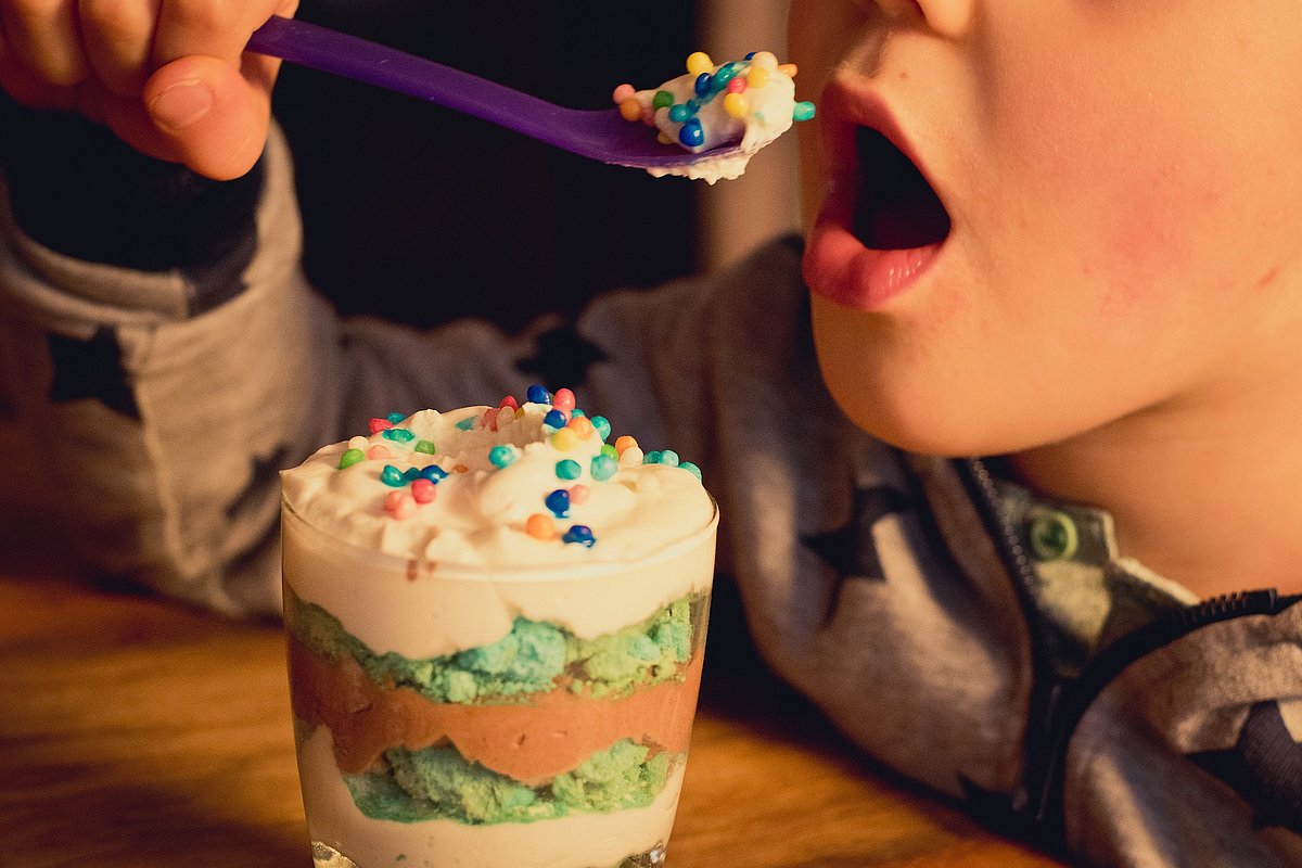Das Foto zeigt in Großaufnahme, wie ein Kind einen Löffel zum geöffneten Mund führt. Der Löffel ist mit einer zuckerreich dekorierten Süßspeise gefüllt, vor dem Kind steht das Dessertglas.
