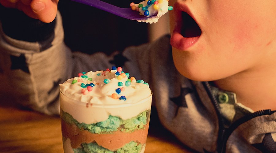 Das Foto zeigt in Großaufnahme, wie ein Kind einen Löffel zum geöffneten Mund führt. Der Löffel ist mit einer zuckerreich dekorierten Süßspeise gefüllt, vor dem Kind steht das Dessertglas.