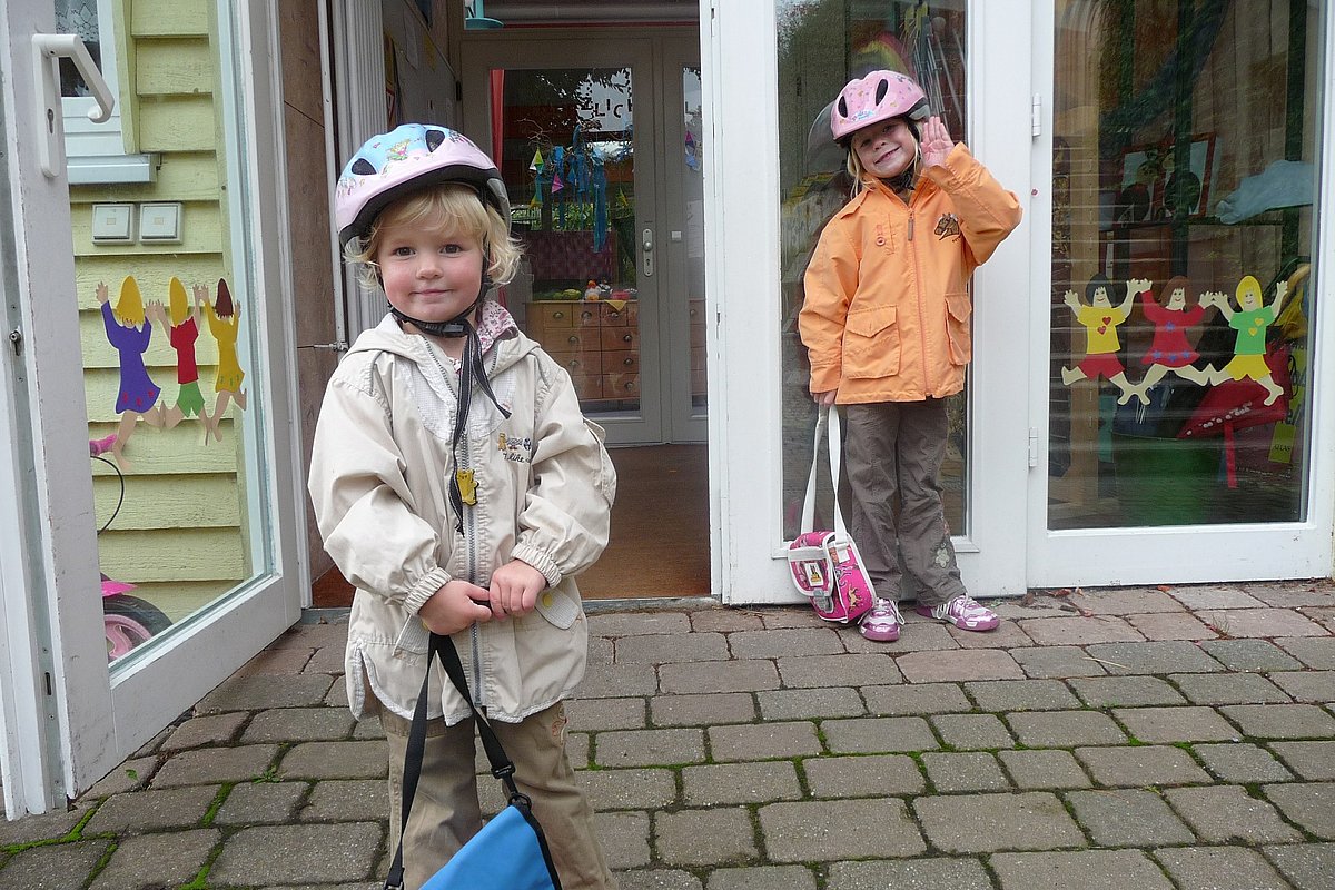 Zwei Kinder mit Fahrradhelm stehen an einer geöffneten Kita-Tür.