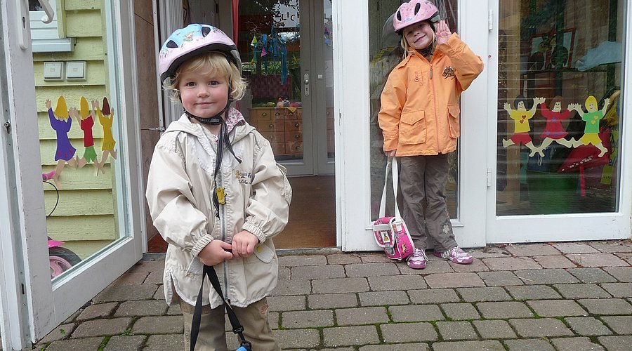 Zwei Kinder mit Fahrradhelm stehen an einer geöffneten Kita-Tür.