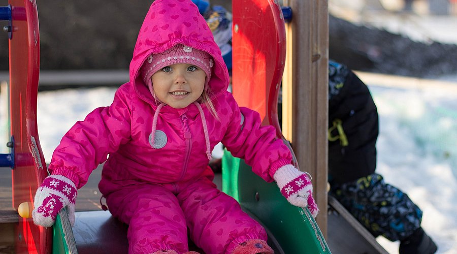 Ein kleines Mädchen im rosa Skianzug sitzt auf einer Rutsche auf einem verschneiten Spielplatz.