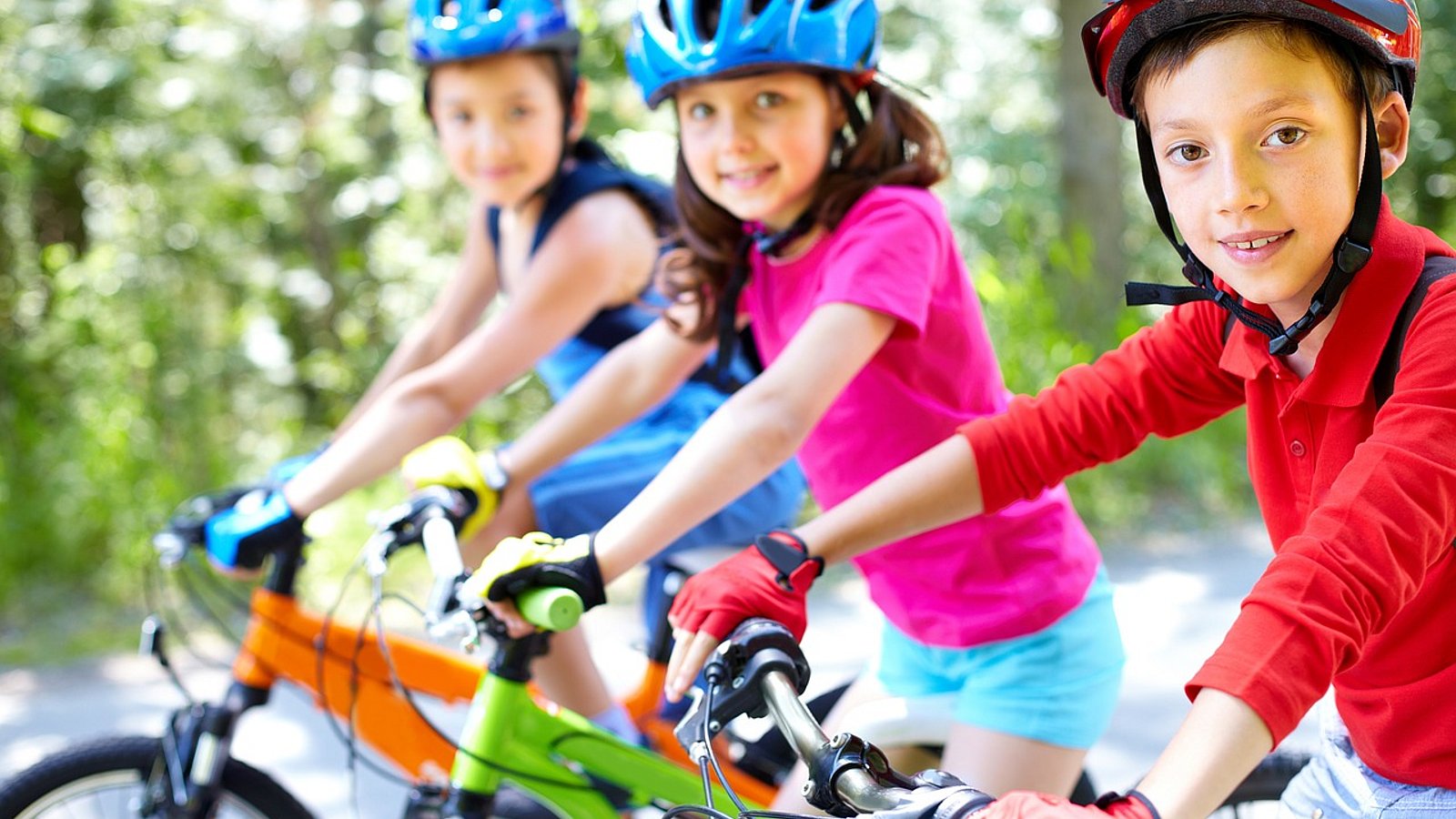 Drei Kinder mit Helm sitzen auf dem Fahrrad und gucken in die Kamera