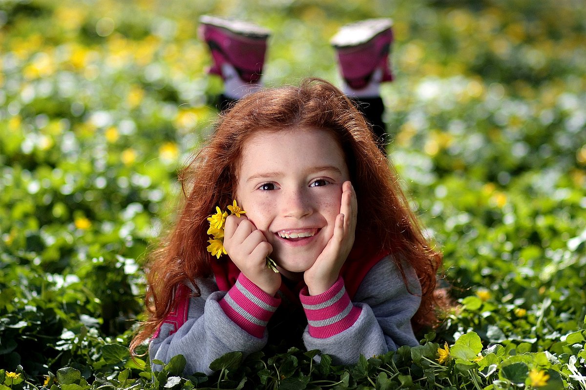 Kindergartenmädchen mit langen roten Haaren liegt bäuchlings auf einer Wiese und lacht mit Blumen in der Hand in die Kamera. 