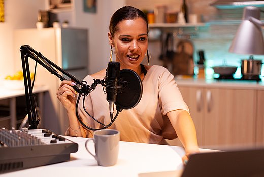 Eine junge Frau mit zurückgesteckten Haaren und großen Ohrringen sitzt in ihrem Home-Podcast-Studio und spricht in ihr Mikrofon.
