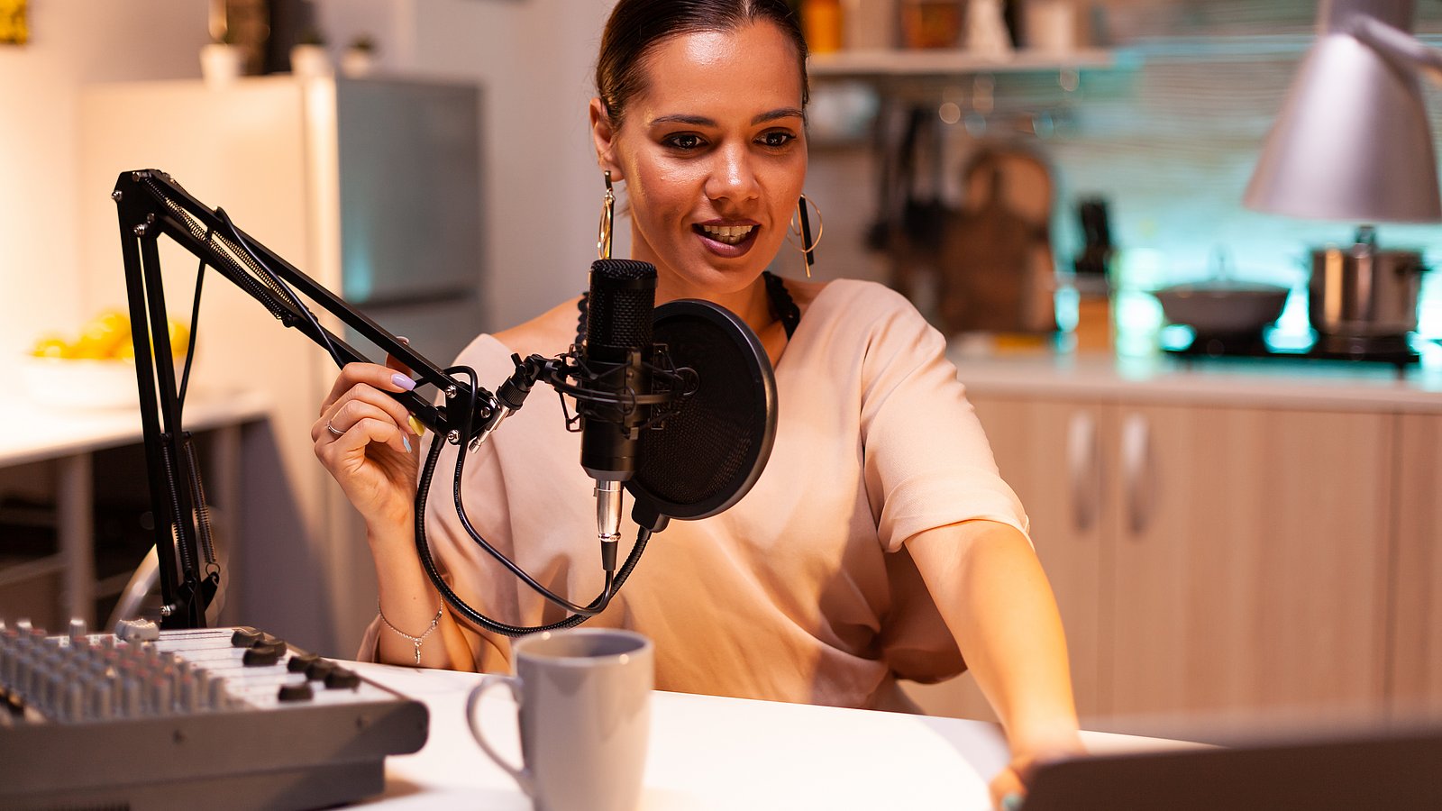 Eine junge Frau mit zurückgesteckten Haaren und großen Ohrringen sitzt in ihrem Home-Podcast-Studio und spricht in ihr Mikrofon.