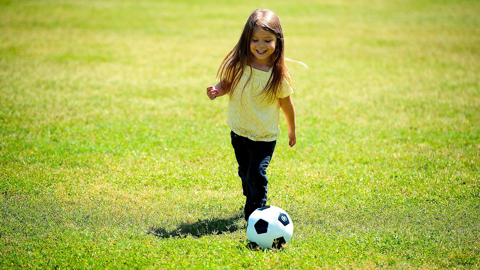 Kleines Mädchen spielt bei Sommerwetter auf dem Rasen mit einem Fußball. 