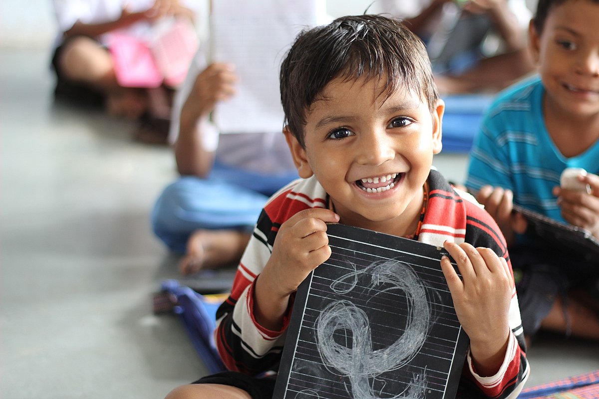 Ein kleiner Schuljunge hält eine Schiefertafel mit einem Buchstaben darauf in der Hand. Er sitzt mit seiner Klasse auf dem Boden.