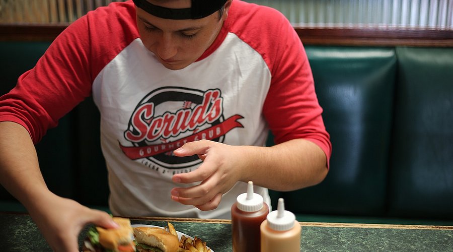Jugendlicher Schüler mit Baseball-Cap sitzt an einem Tisch und isst einen Burger. 