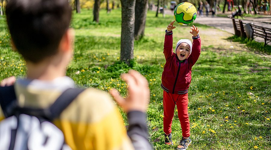 Ein Junge im Kitaalter wirft auf einem Rasen in einem Park einen gelben Ball einem älteren Jungen zu. Der kleinere Junge lacht fröhlich, der ältere Junge zeigt sich von der Rückenansicht, er trägt einen Rucksack und hält die Hände zum Fangen bereit. . 