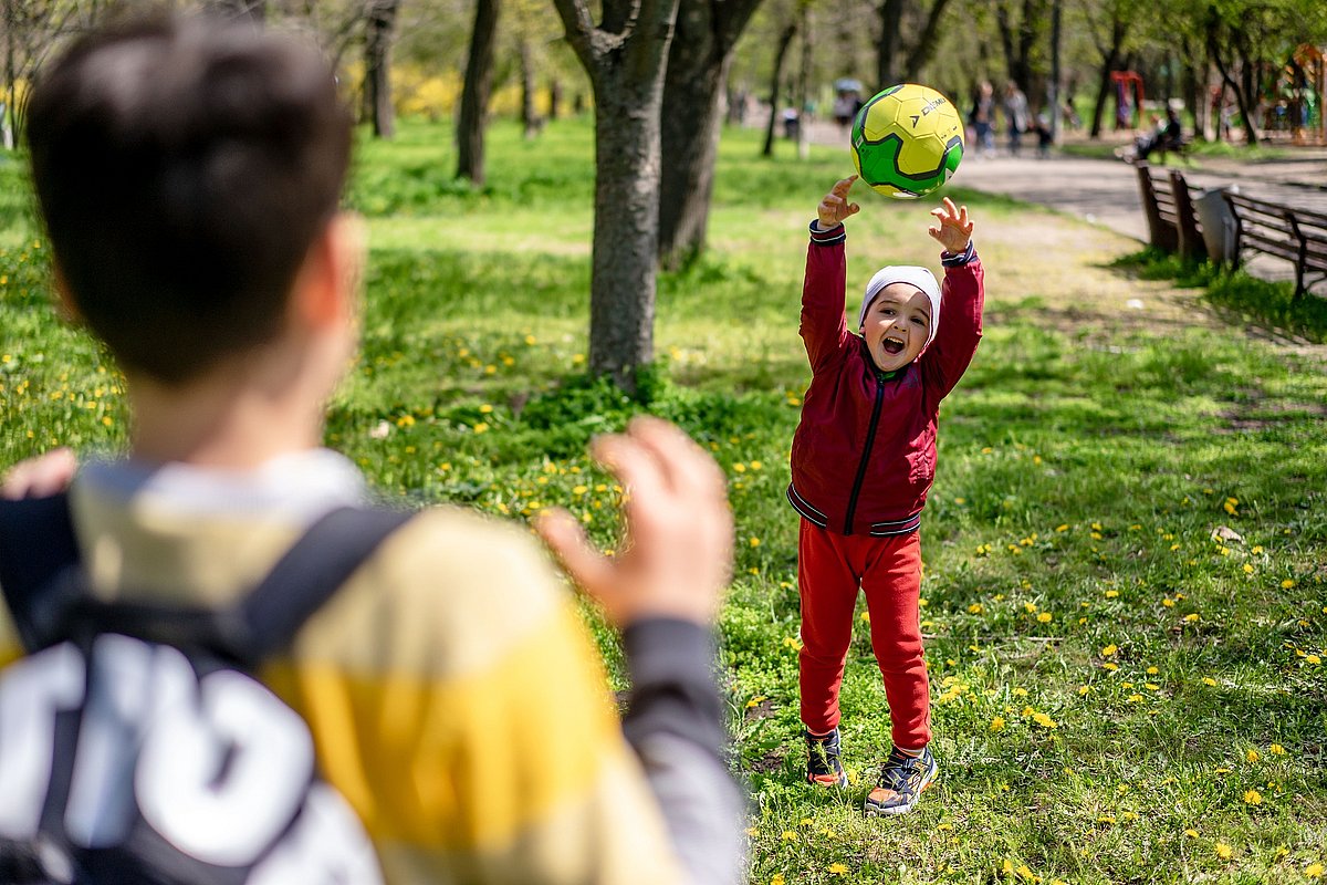 Ein Junge im Kitaalter wirft auf einem Rasen in einem Park einen gelben Ball einem älteren Jungen zu. Der kleinere Junge lacht fröhlich, der ältere Junge zeigt sich von der Rückenansicht, er trägt einen Rucksack und hält die Hände zum Fangen bereit. . 
