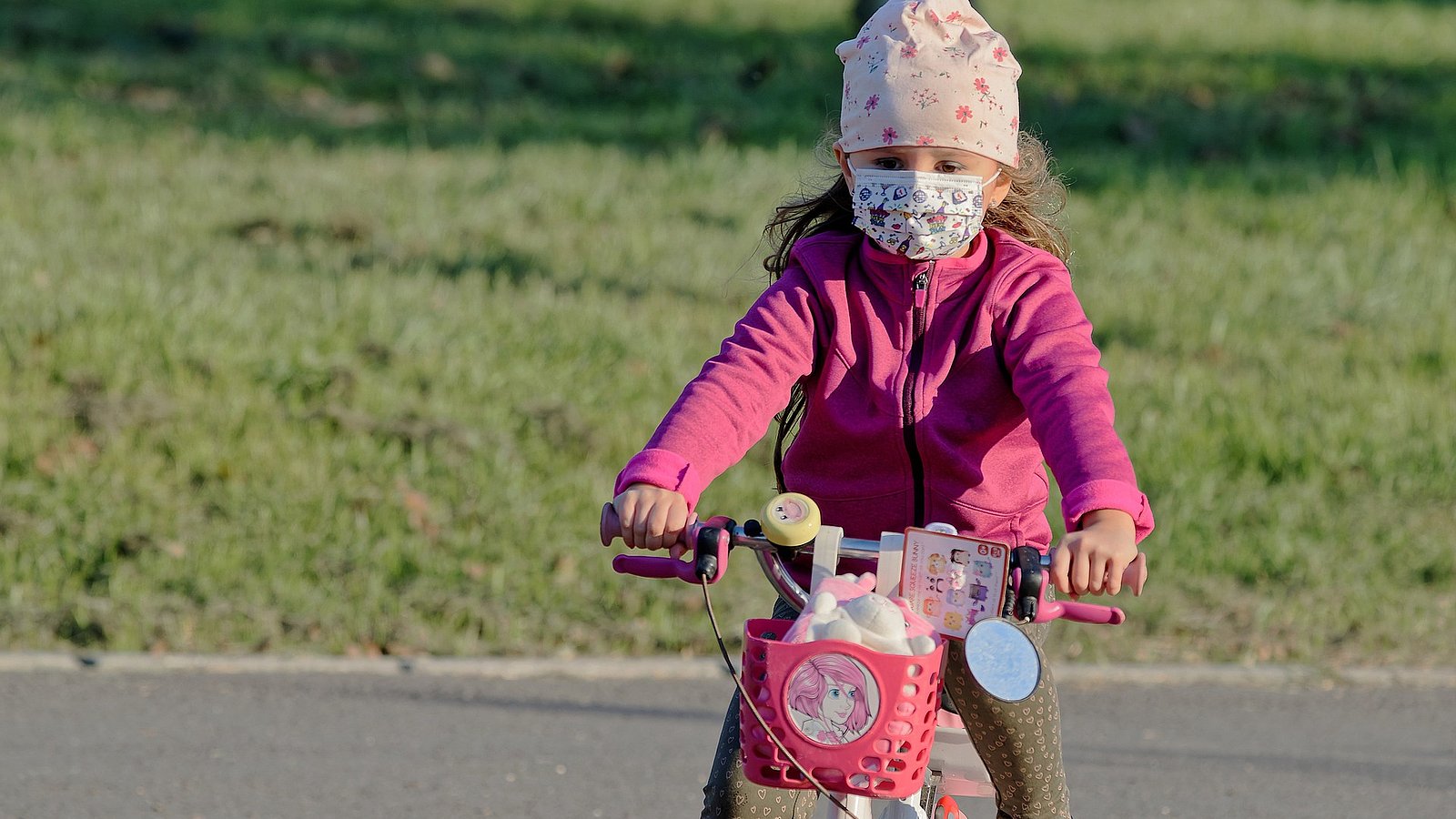 Kleines Mädchen hat pandemiebedingt eine Maske auf und fährt auf ihrem Kinderfahrrad. 