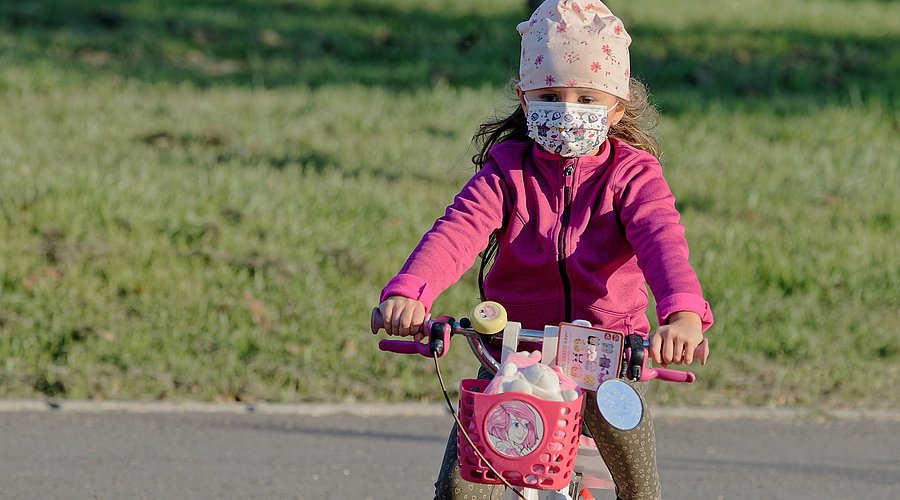 Kleines Mädchen hat pandemiebedingt eine Maske auf und fährt auf ihrem Kinderfahrrad. 