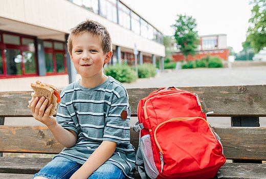Ein Schuljunge mit kurzen blonden Stoppelhaaren sitzt verschmitzt lachend auf einer Holzbank vor seiner Schule und hält ein angebissenes Butterbrot in der Hand. Neben sich steht sein roter Schulrucksack.
