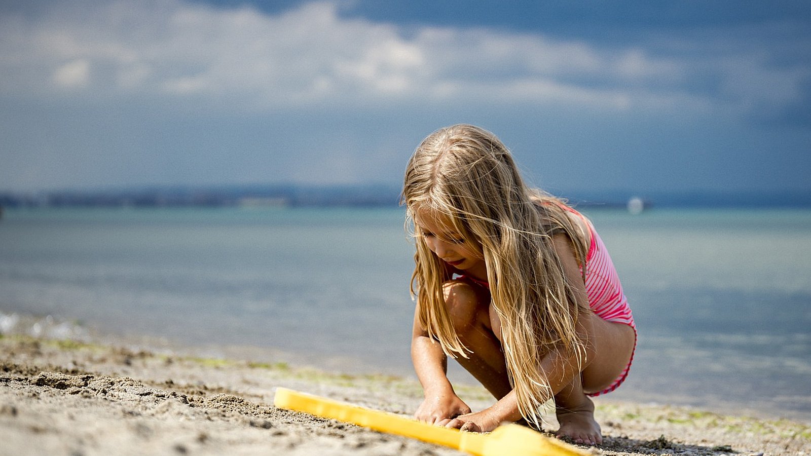 Kleines Mädchen mit langen blonden Haaren spielt im Badeanzug an einem Sandstrand. 
