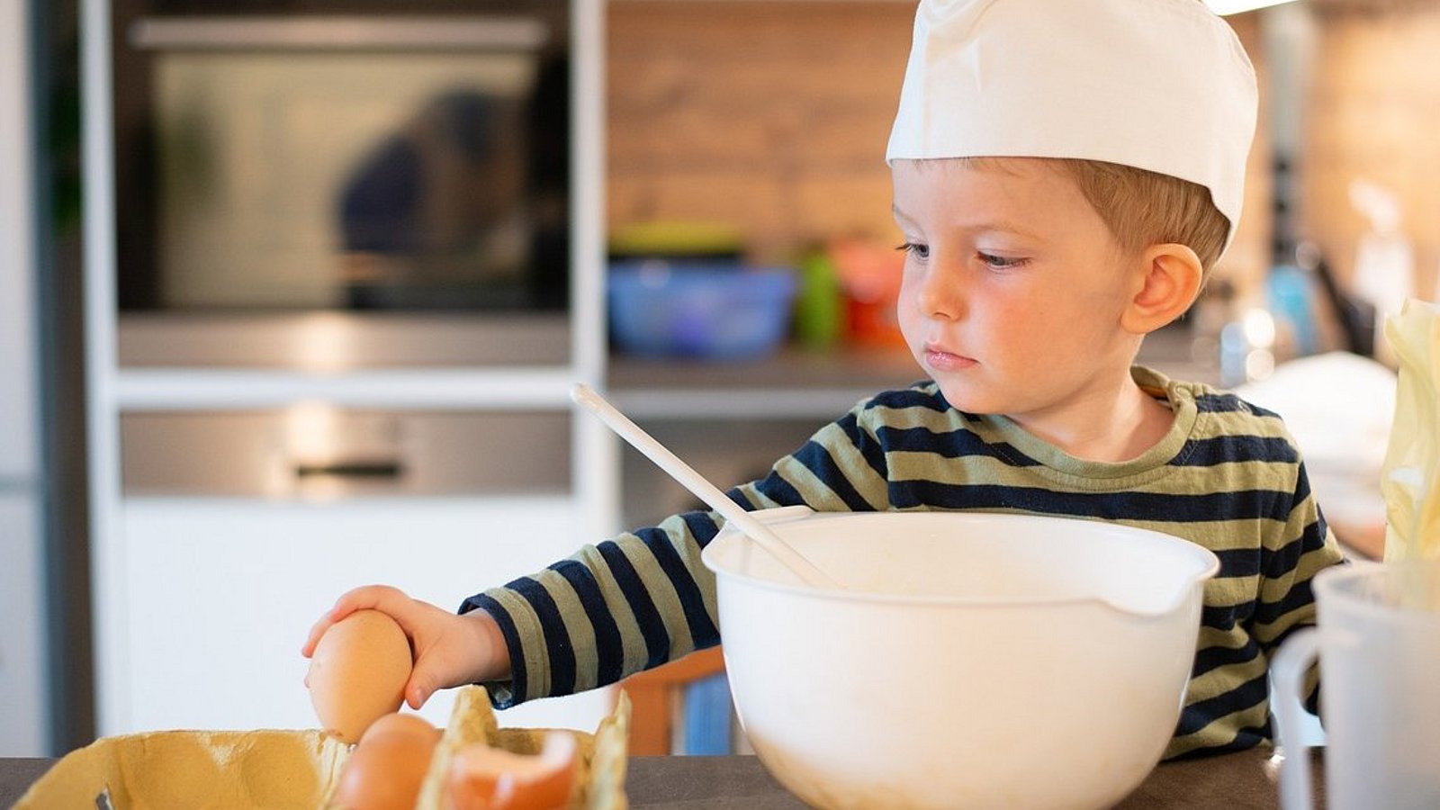 Ein Junge im Kindergartenalter steht an einem Küchentisch und hat eine Rührschüssel vor sich. Er nimmt ein rohes Ei aus einem Eierkarton. 