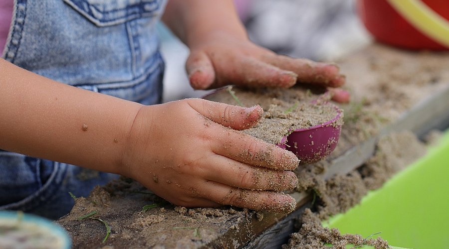 Ein kleines Kind spielt am Sandkastenrand und drückt Sand in ein Förmchen. Das Bild zeigt vor allem die Hände des Kindes, Sand und Sandform..
