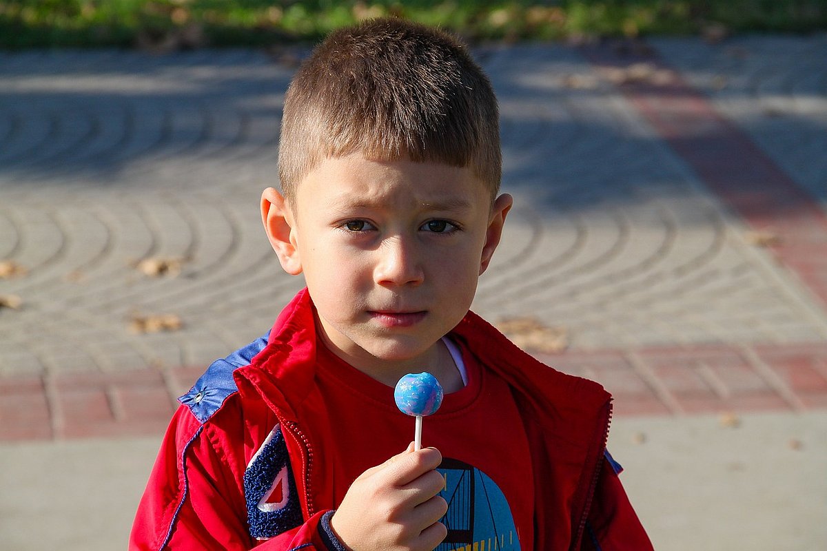Ein kleiner Junge mit einer roten Jacke hält einen künstlich blauen Lolli in der Hand. 