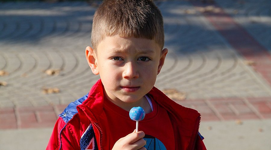 Ein kleiner Junge mit einer roten Jacke hält einen künstlich blauen Lolli in der Hand. 