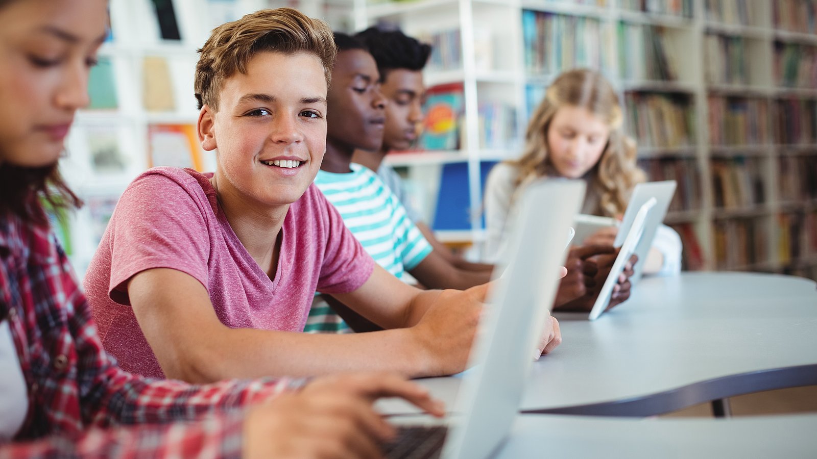 Jugendliche Schüler*innen sitzen in einer Schulbibliothek am Laptop. Ein Junge schaut lächelnd in die Kamera, alle anderen konzentrieren sich auf ihre Arbeit. 