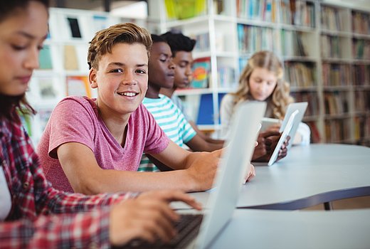 Jugendliche Schüler*innen sitzen in einer Schulbibliothek am Laptop. Ein Junge schaut lächelnd in die Kamera, alle anderen konzentrieren sich auf ihre Arbeit. 