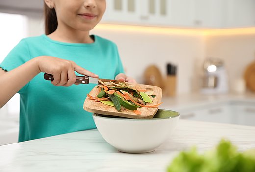 Ein junges Mädchen steht in einer überwiegend weißen Küche an einem Arbeitstisch. Sie hält ein Holzbrettchen mit Abfallresten vom Gemüseputzen in der Hand, die sie mit einem Messer in eine Schüssel schiebt.