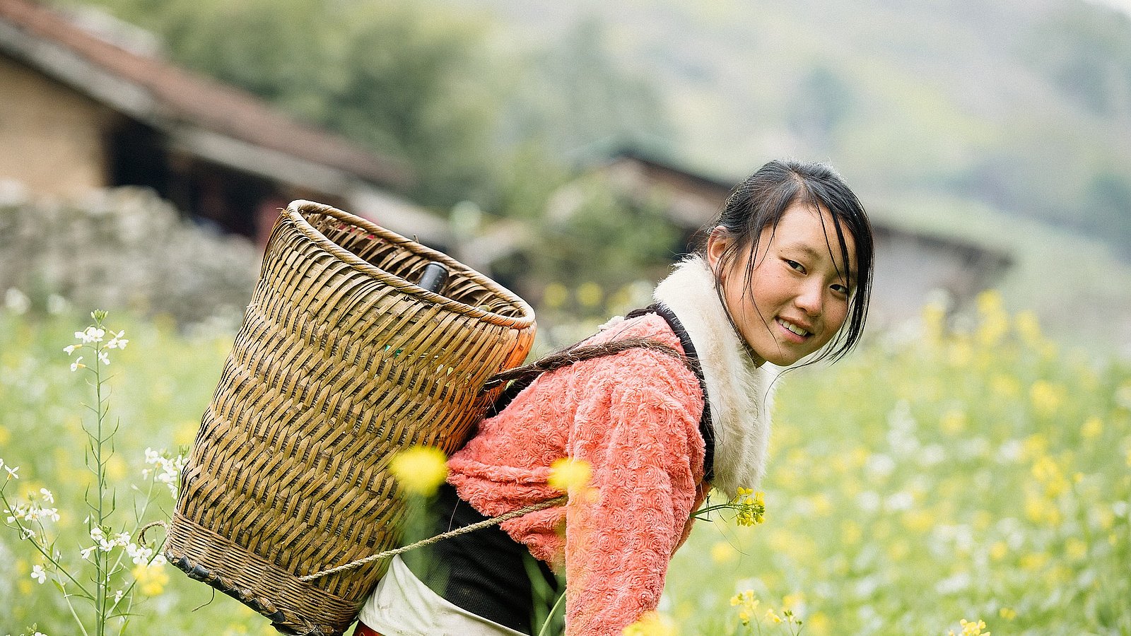 Ein junges Mädchen hält einen Erntekorb auf ihrem Rücken und steht gebeugt in einem Feld. Sie lächelt in die Kamera.