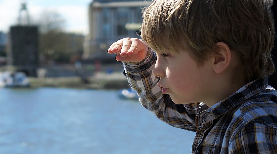 Ein Junge schaut in die Ferne und legt dabei die Hand über die Augen. Im Hintergrund ein See oder das Meer.