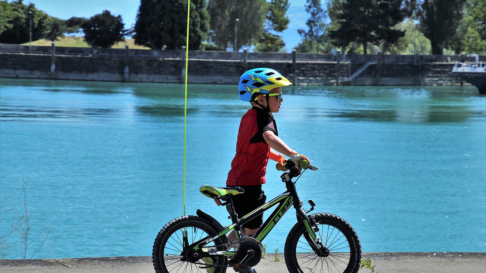 Ein Junge im Kitaalter steht neben seinem Fahrrad an einem See. Er trägt einen Helm, das Fahrrad hat einen Sicherheitswimpel.