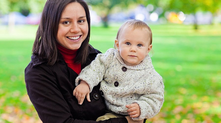 Eine junge Mutter steht in einem herbstlichen Park und hält ihr Baby auf dem Arm. 