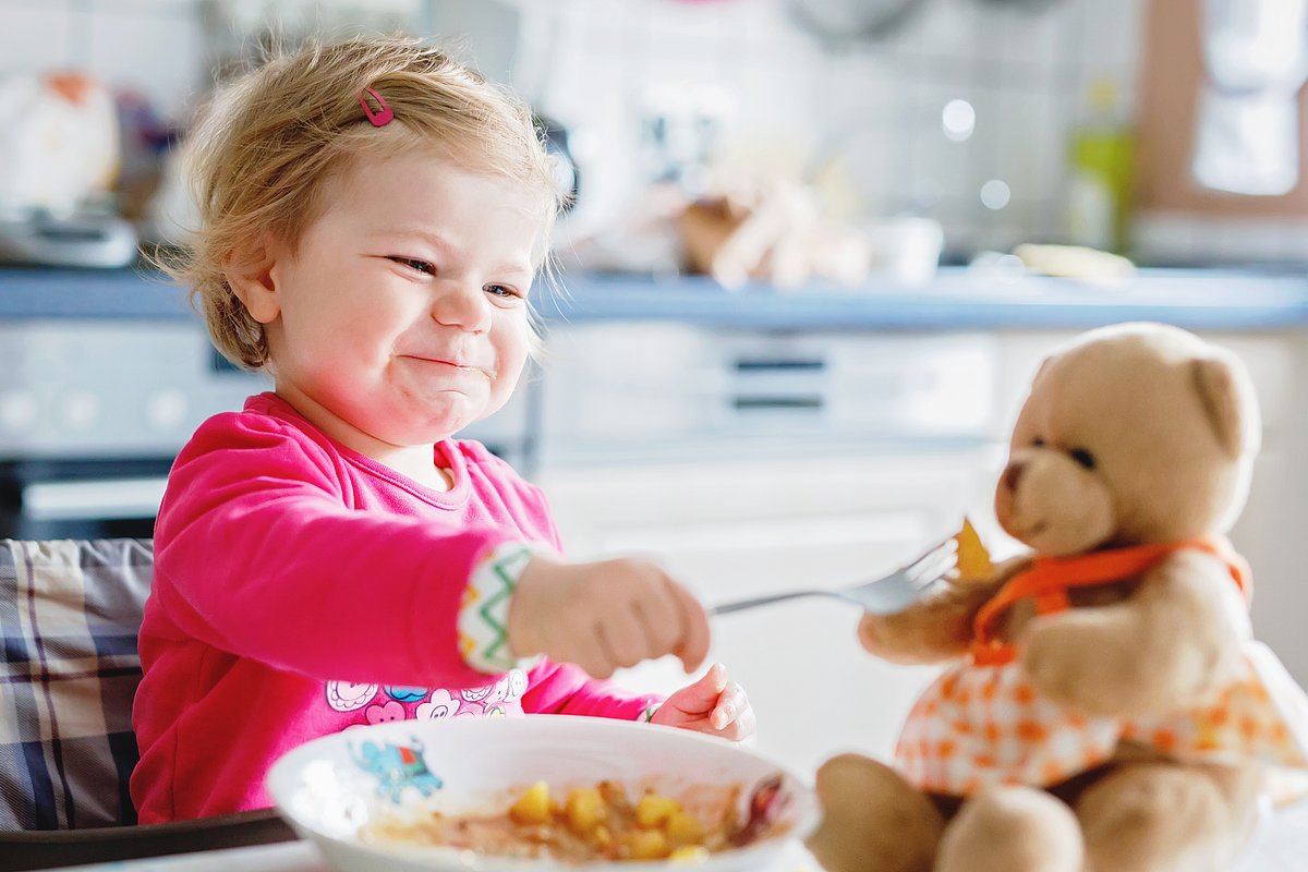 Kleines Mädchen sitzt im Hochstuhl am Tisch und füttert ihren Teddybären. 