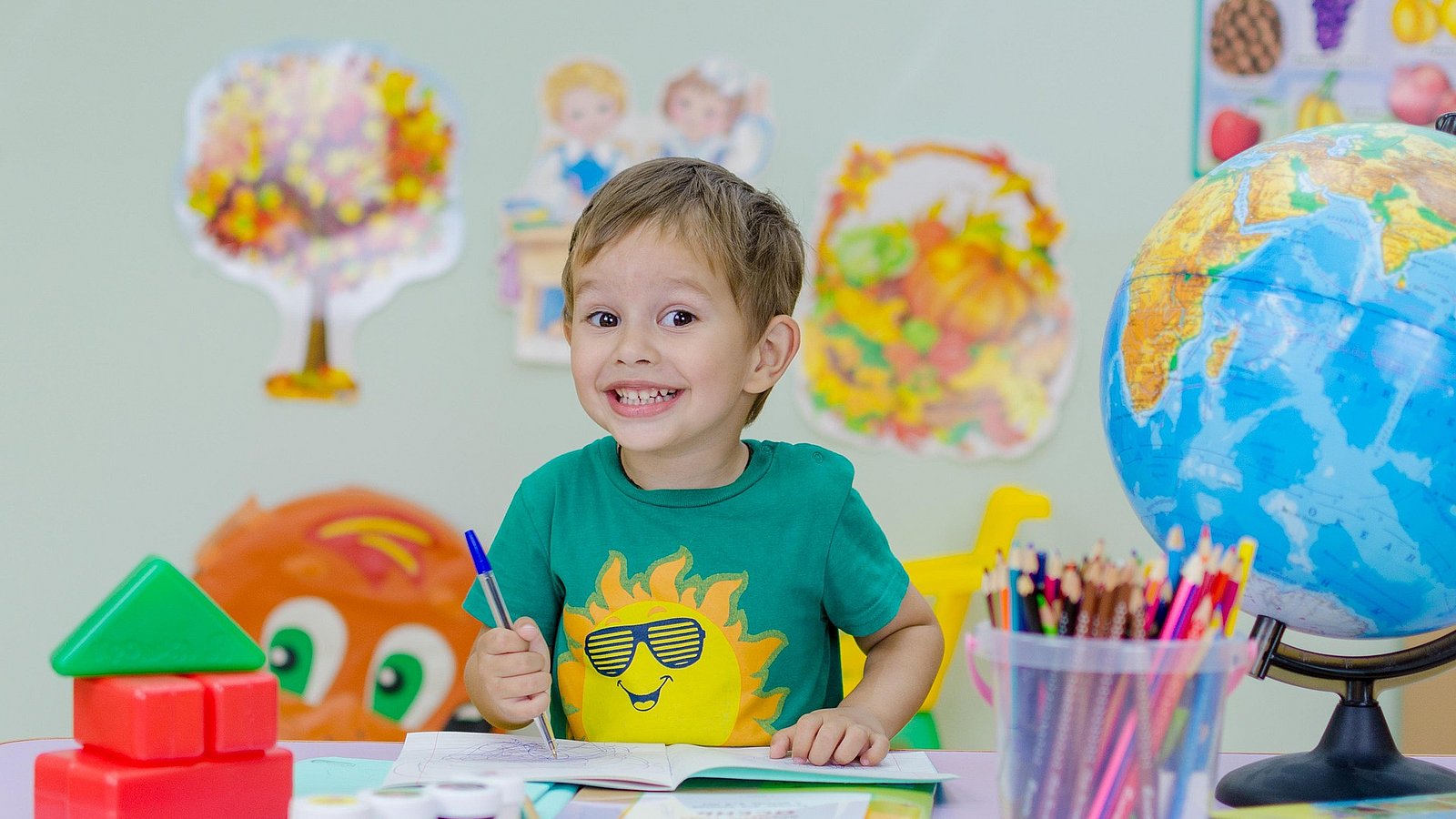 Kleiner Schuljunge sitzt am Schreibtisch, vor ihm ein aufgeschlagenes Lehrbuch, bunte Stifte und ein Globuls. Er lacht erwartungsvoll in die Kamera.