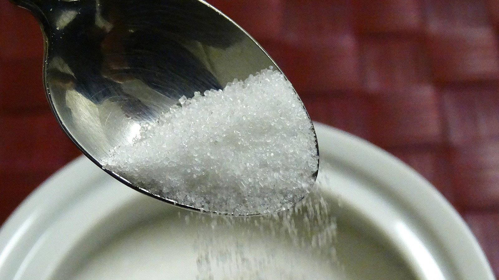 Von einem Esslöffel rieselt Zucker in eine Zuckerdose.