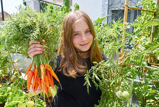 Ein jugendliches Schulmädchen mit langen glatten Haaren hält in einem Garten inmitten von Tomatenstauden einen Bund Möhren in der Hand. Sie trägt eine Kette sehr hoch am Hals.