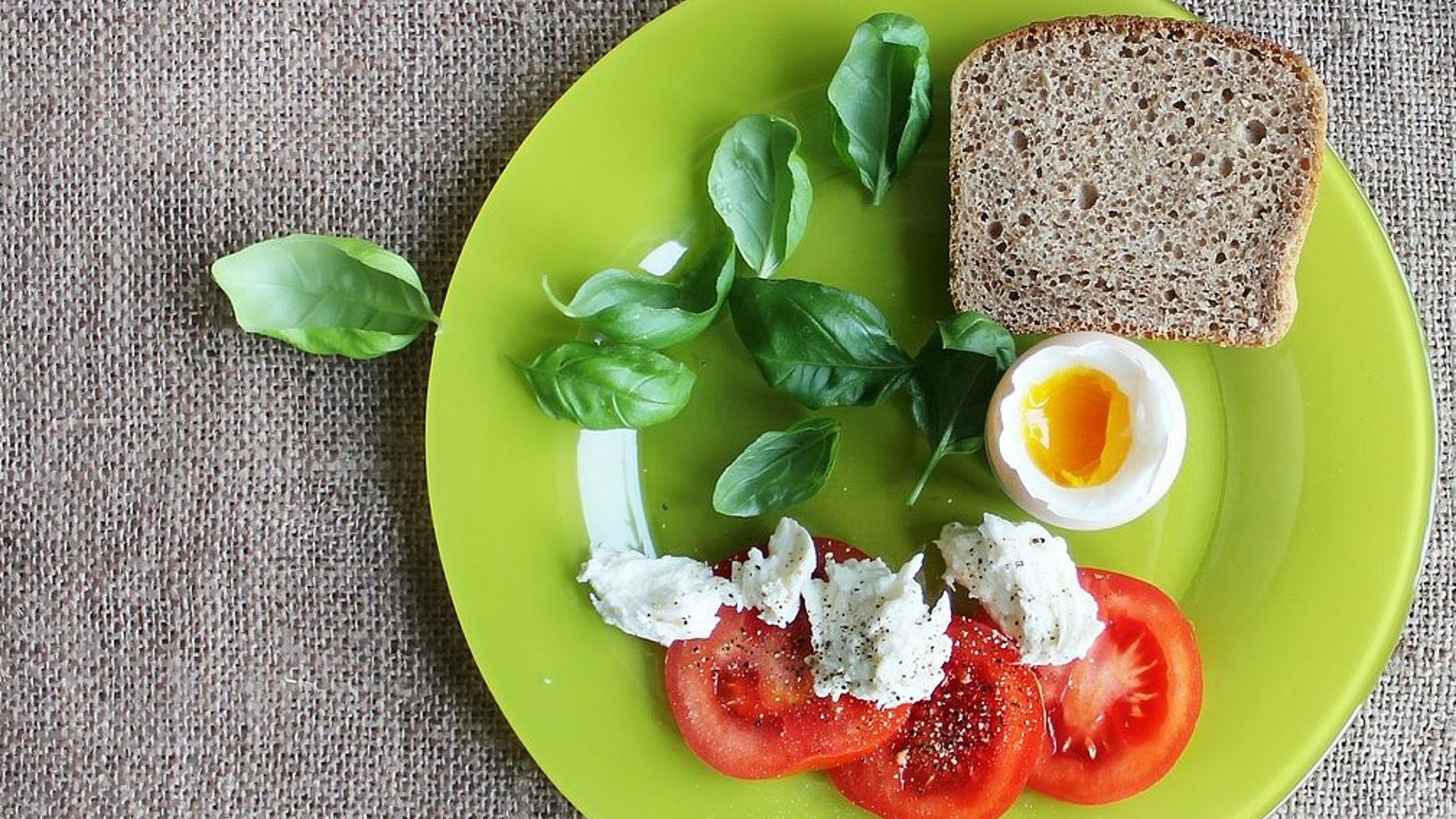 Auf einem grünen Teller sind drei Tomatenscheiben mit Feta-Käse, Basilikumblättern und einer Scheibe Vollkornbrot angerichtet. In der Mitte liegt ein halbes Ei. 