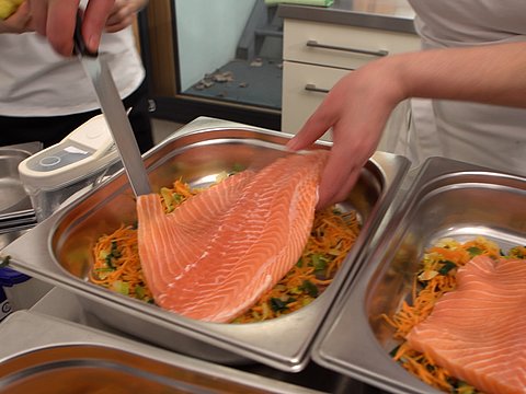 In einer Kita-Frischküche in Wiesbaden legt eine Küchenkraft Lachs auf eine Gemüsebett.