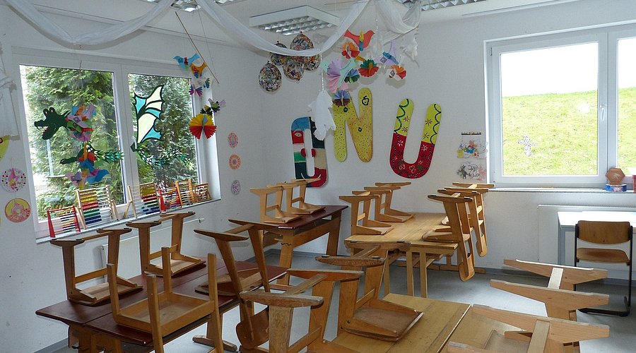 Ein leeres Klassenzimmer einer Grundschule mit, die Stühle sind hochgestellt. An den Wänden hängen bunte Bilder der Schülerinnen und Schüler.