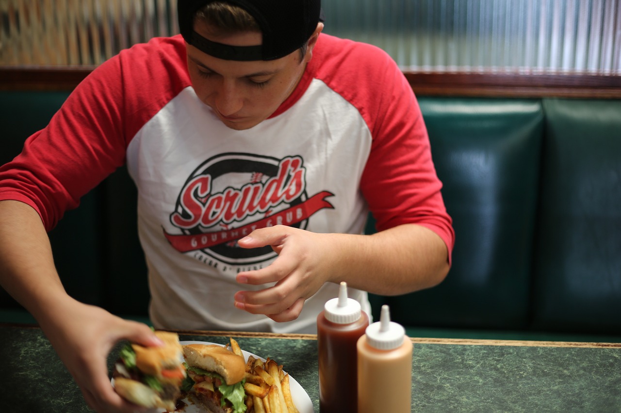 Jugendlicher Schüler mit Baseball-Cap sitzt an einem Tisch und isst einen Burger. 