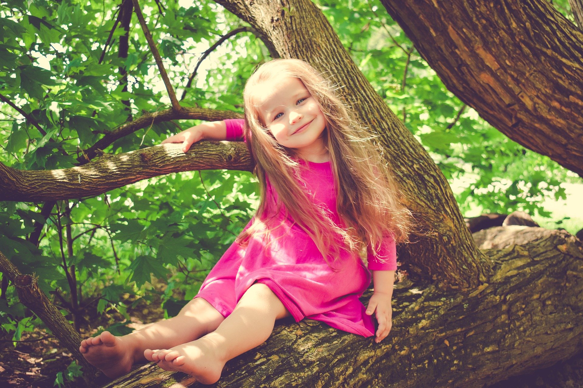 Kleines Mädchen sitzt barfuß auf einem Baumstamm.